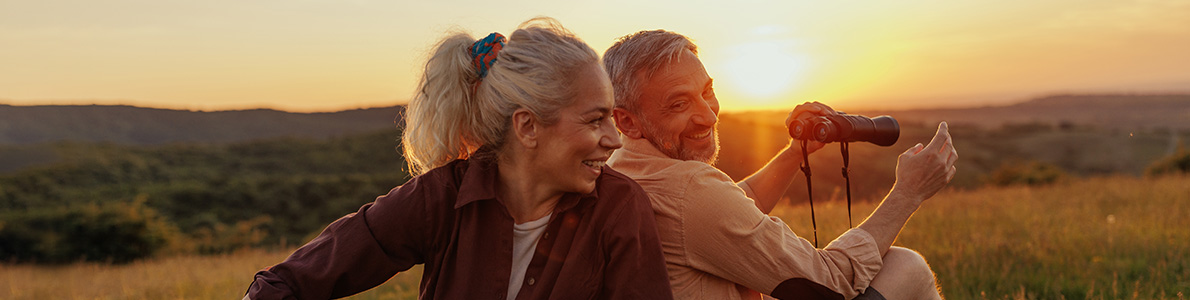 outdoorsy couple smiling and sitting back to back on a grass landscape with binoculars