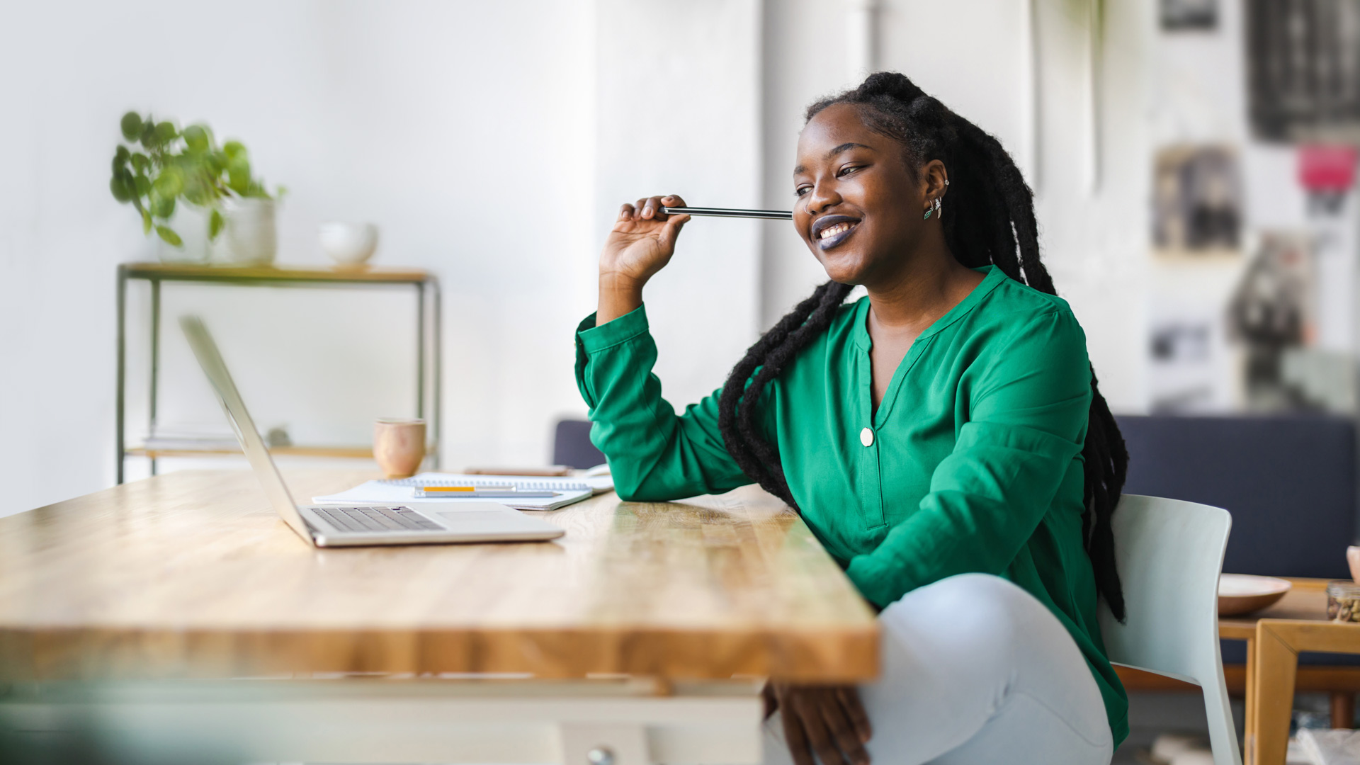 A Black woman with long locs and a bright green shirt sits at a wooden desk as she smiles and types on a laptop.