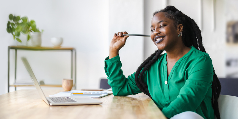 A Black woman with long locs and a bright green shirt sits at a wooden desk as she smiles and types on a laptop.
