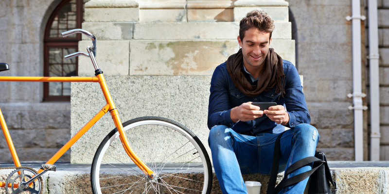 A white man wearing a blue shirt and brown scarf sits on a curb next to his yellow bike. He smiles while looking at his phone.