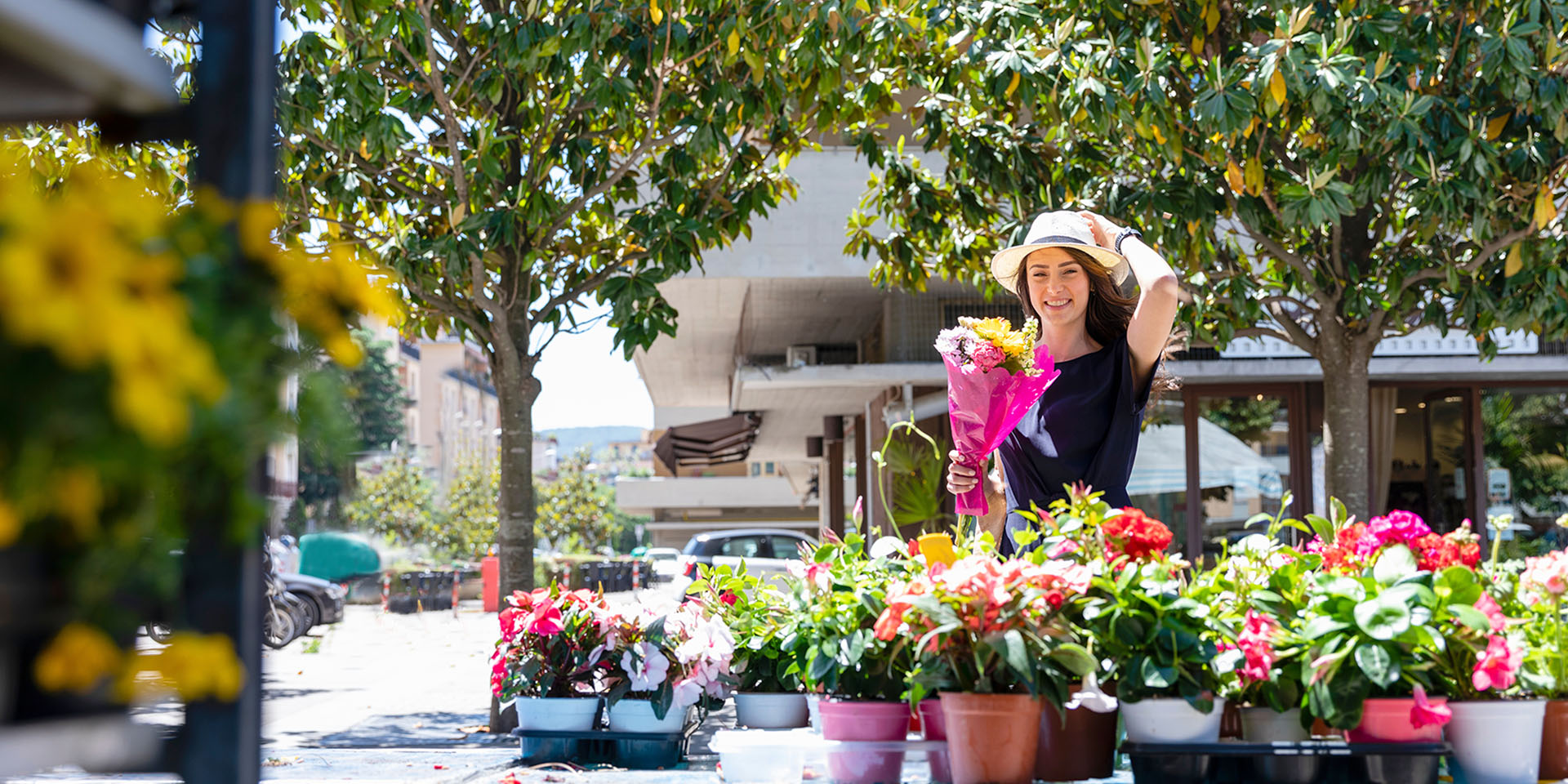 A white woman with long brown hair in a blue dress and sun hat smiles while holding flowers at an outdoor market.