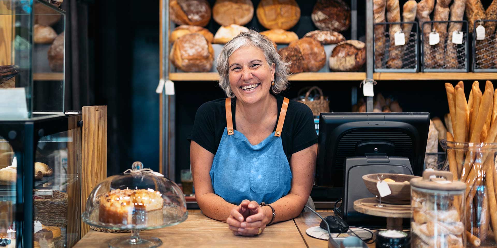 A woman smiles as she leans over the counter of her bakery, smiling at you.