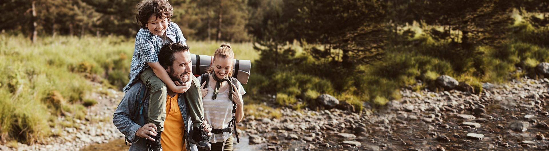 A mom and dad hike across a shallow creek in a forested landscape with their son on the dad's shoulders.