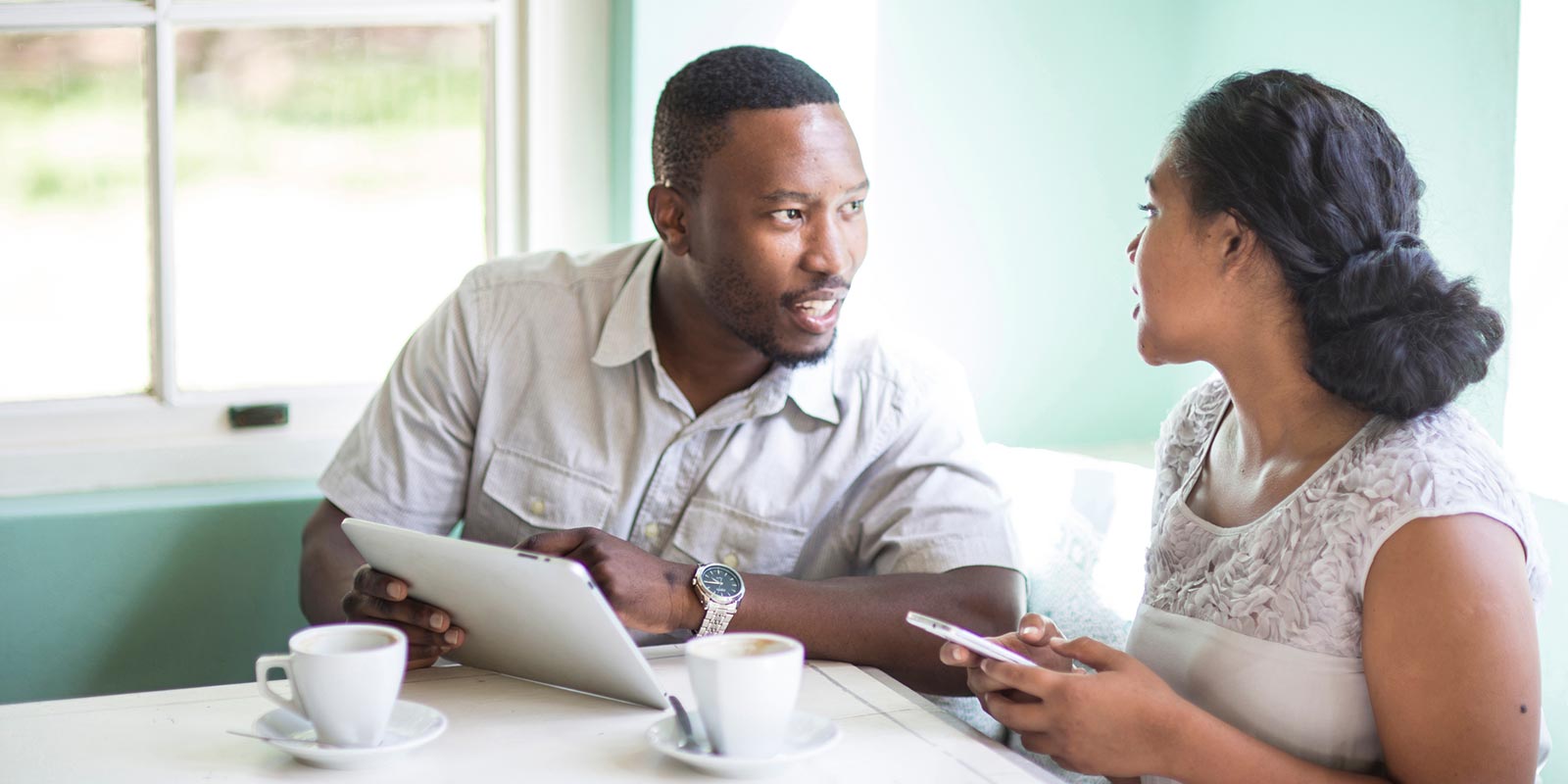 A Black man and woman sit at a table discussing paperwork over coffee in a cafe.