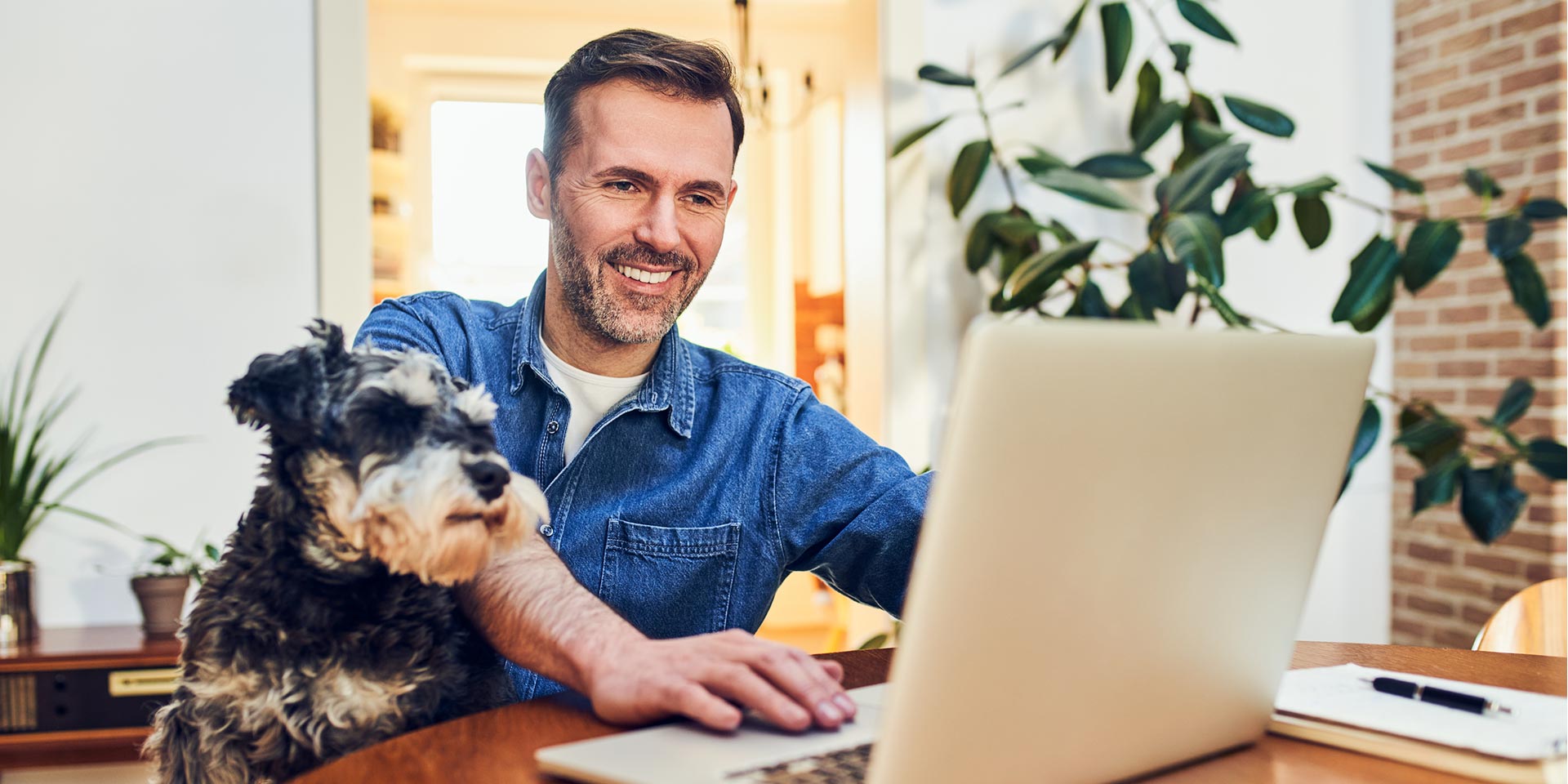 man at a table on his laptop smiling with his dog next to him