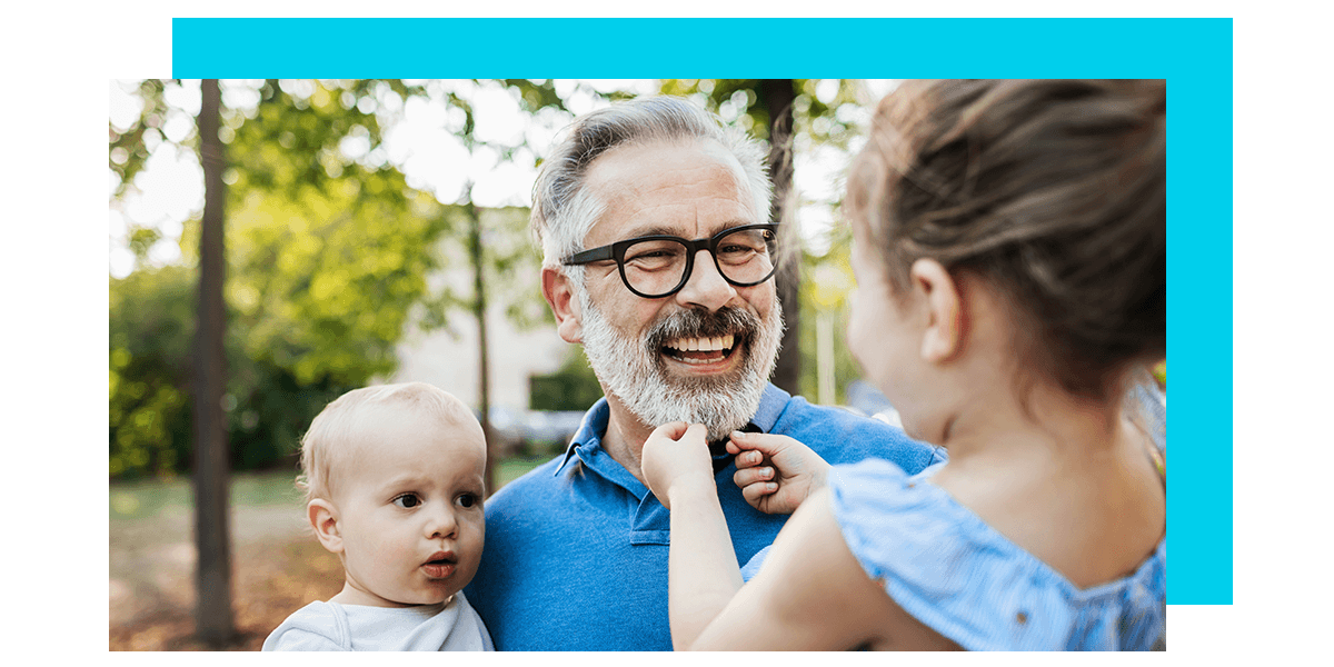 smiling older man holding two young children outside in nature.