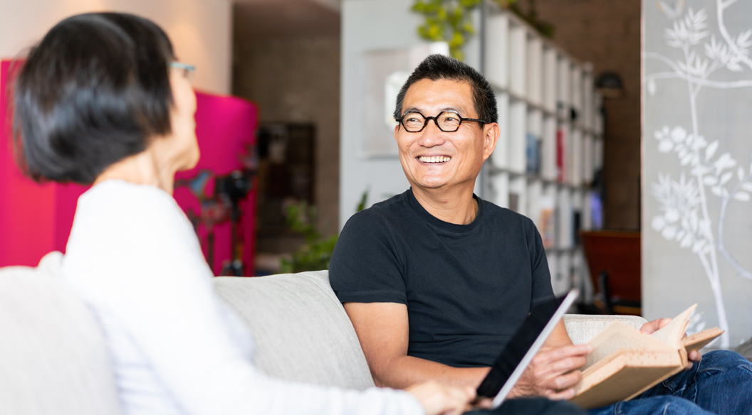 Man and woman smiling at each other on the couch with a book and a laptop.