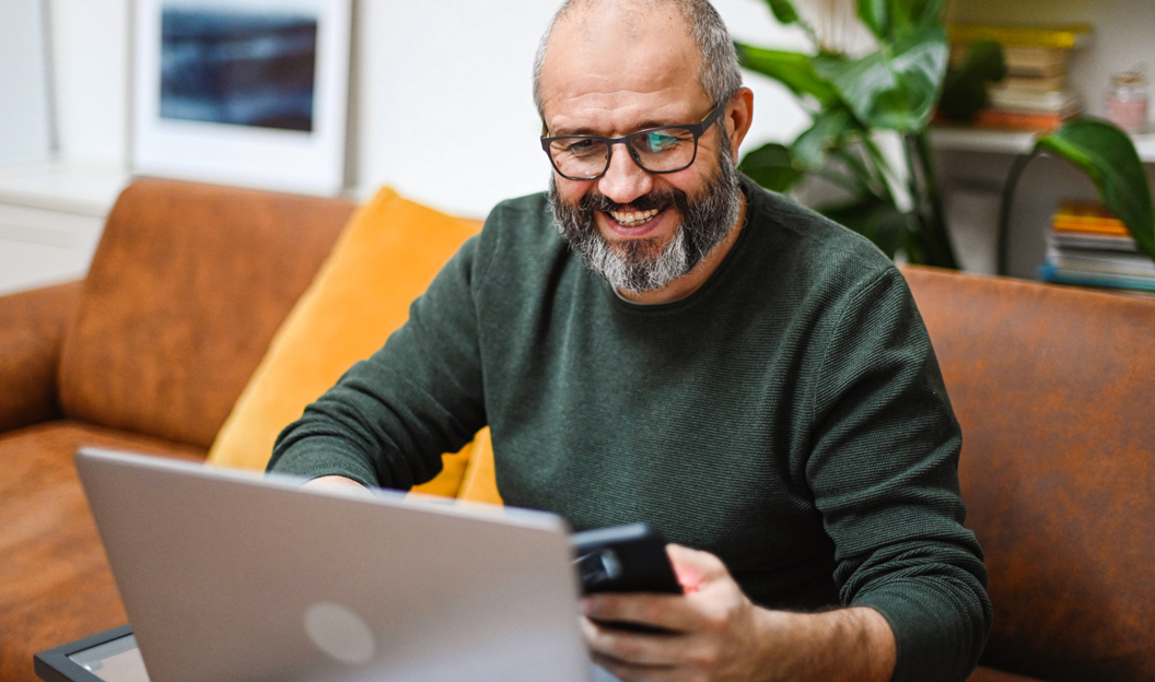 man on couch with his laptop smiling down at his phone.