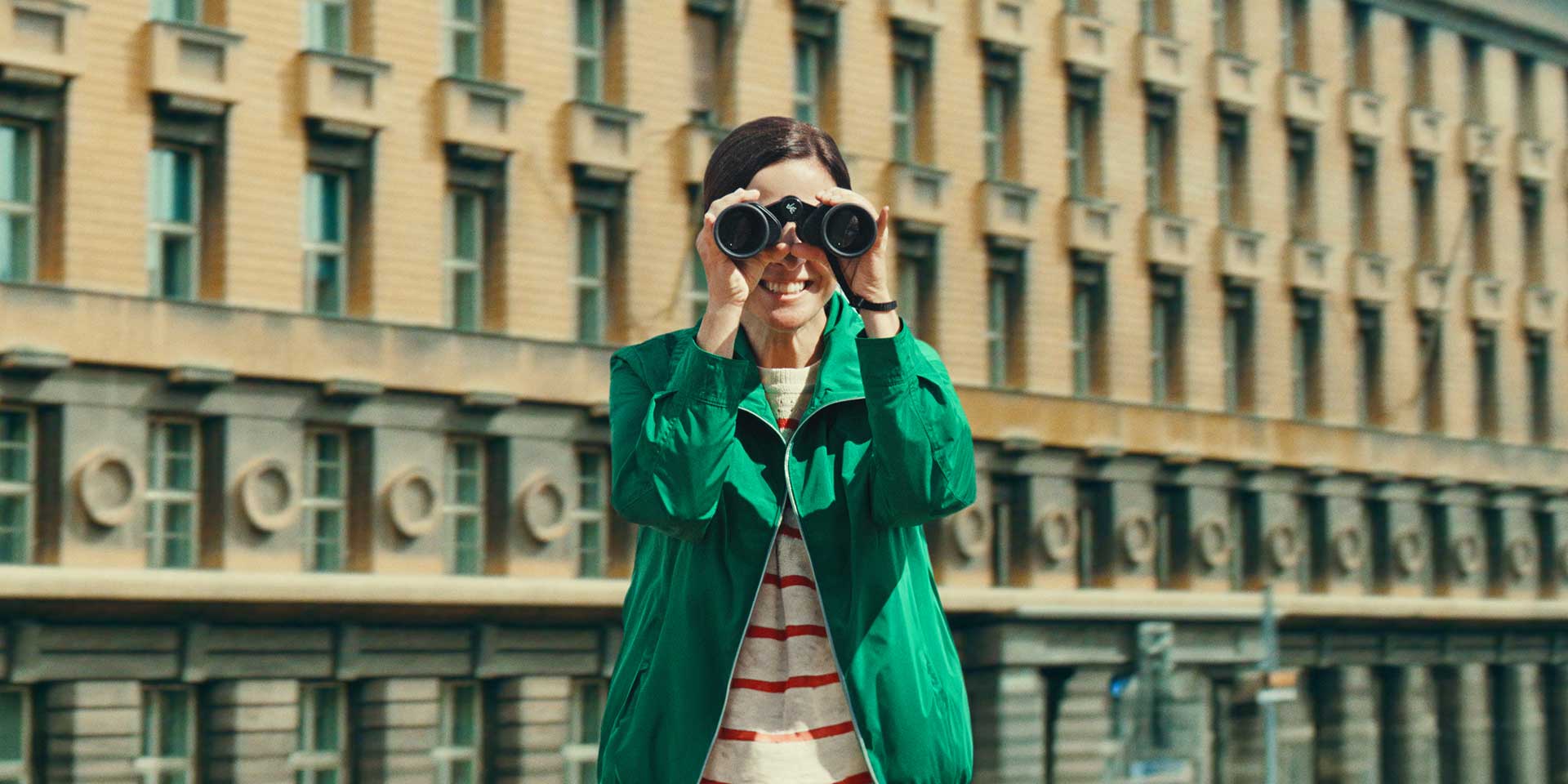 A woman in a green coat stands in front of a city building. She looks through a pair of binoculars in your direction, smiling.