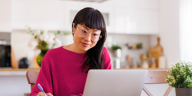 woman sitting at her kitchen table looking at her laptop while writing with a pen