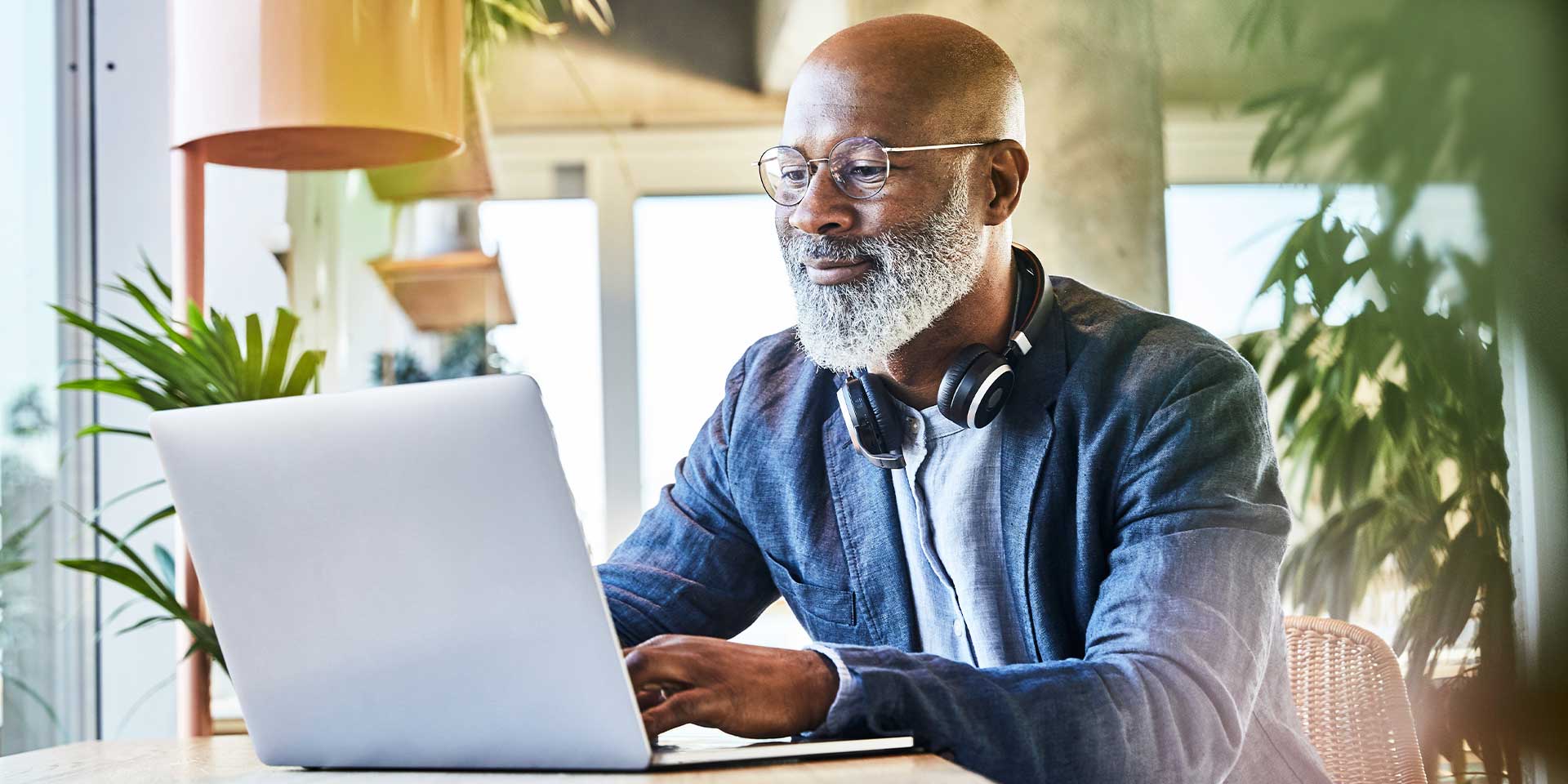 An older Black man wearing glasses and a denim shirt sits in a plant shop while working at his laptop.