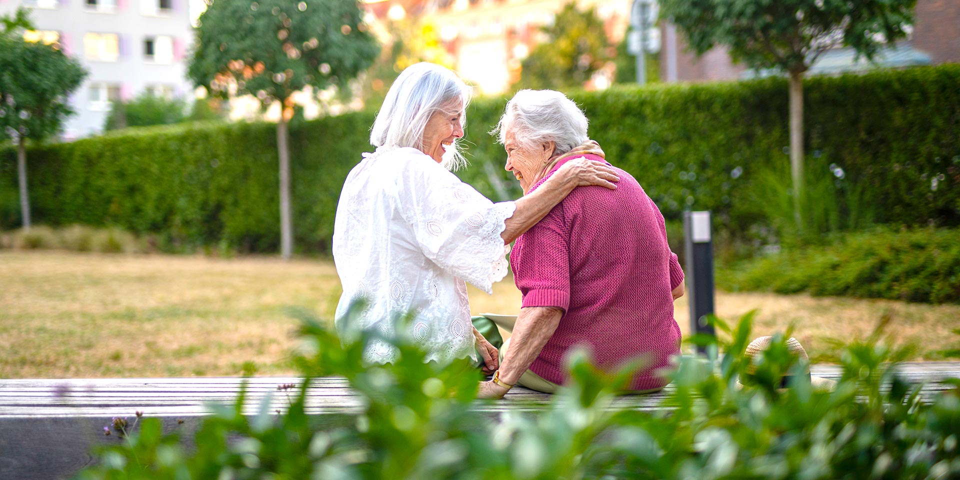 Two older White women sit on a park bench hugging and laughing.