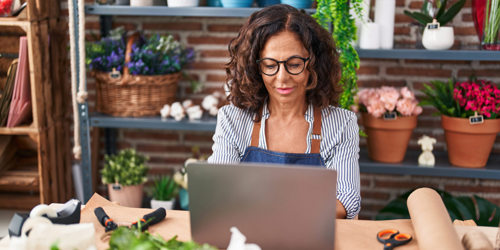 A middle-aged female florist sits at her desk inside her flowershop, working on her computer.