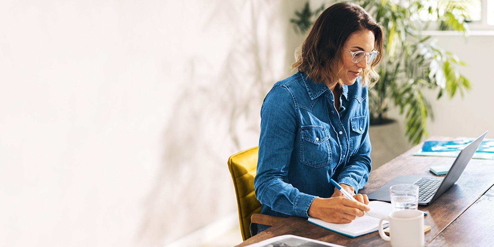 Woman at a desk smiling at her laptop while writing in a notepad
