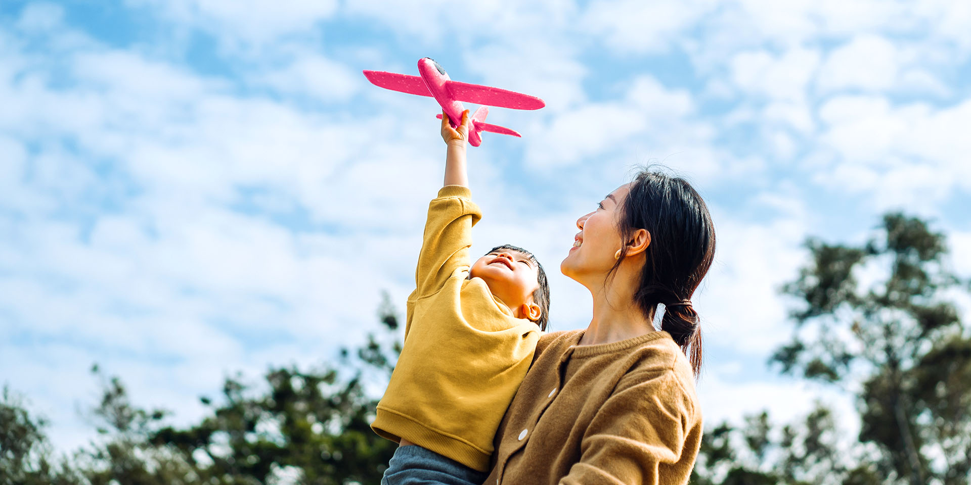 Mom holding her son outside while he holds up a red toy plane 