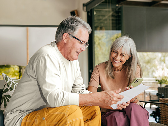Older couple on the couch smiling while holding a few papers and a cell phone as if planning