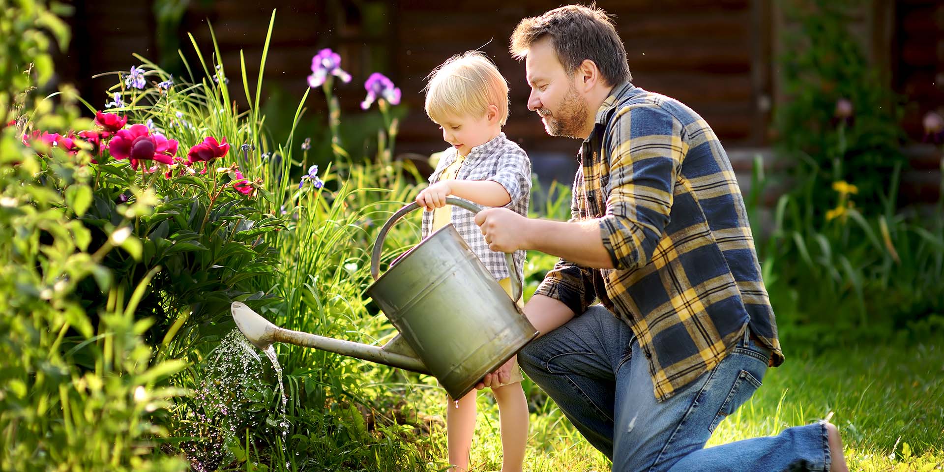 dad kneeling down with son with both of their hands on the handle of a watering can in front of a garden