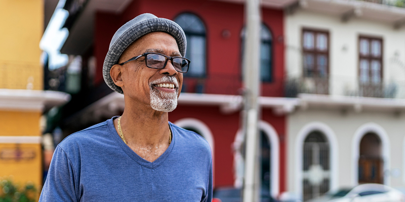older man with a hat and sunglasses smiling outside in front of a cute building