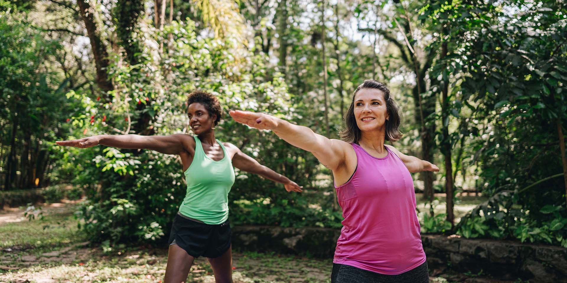 two women outside in a forest clearing doing yoga together, reaching their arms wide in warrior two position