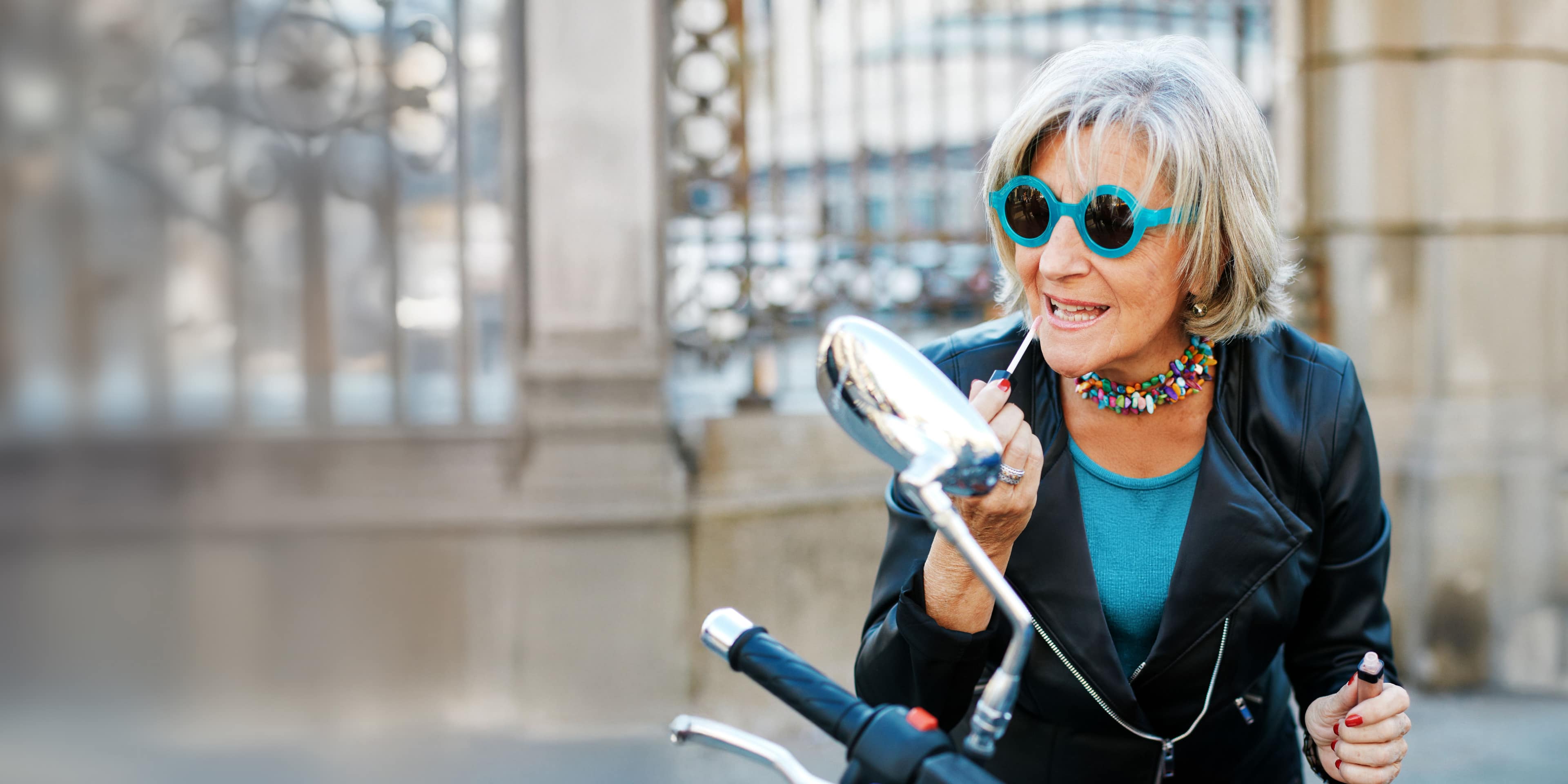 A vibrant retiree in stylish sunglasses applies lip gloss in the side-view mirror of her parked motorcycle.