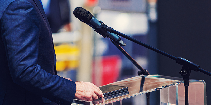 Male speaker on the stage wearing suit at media press conference.