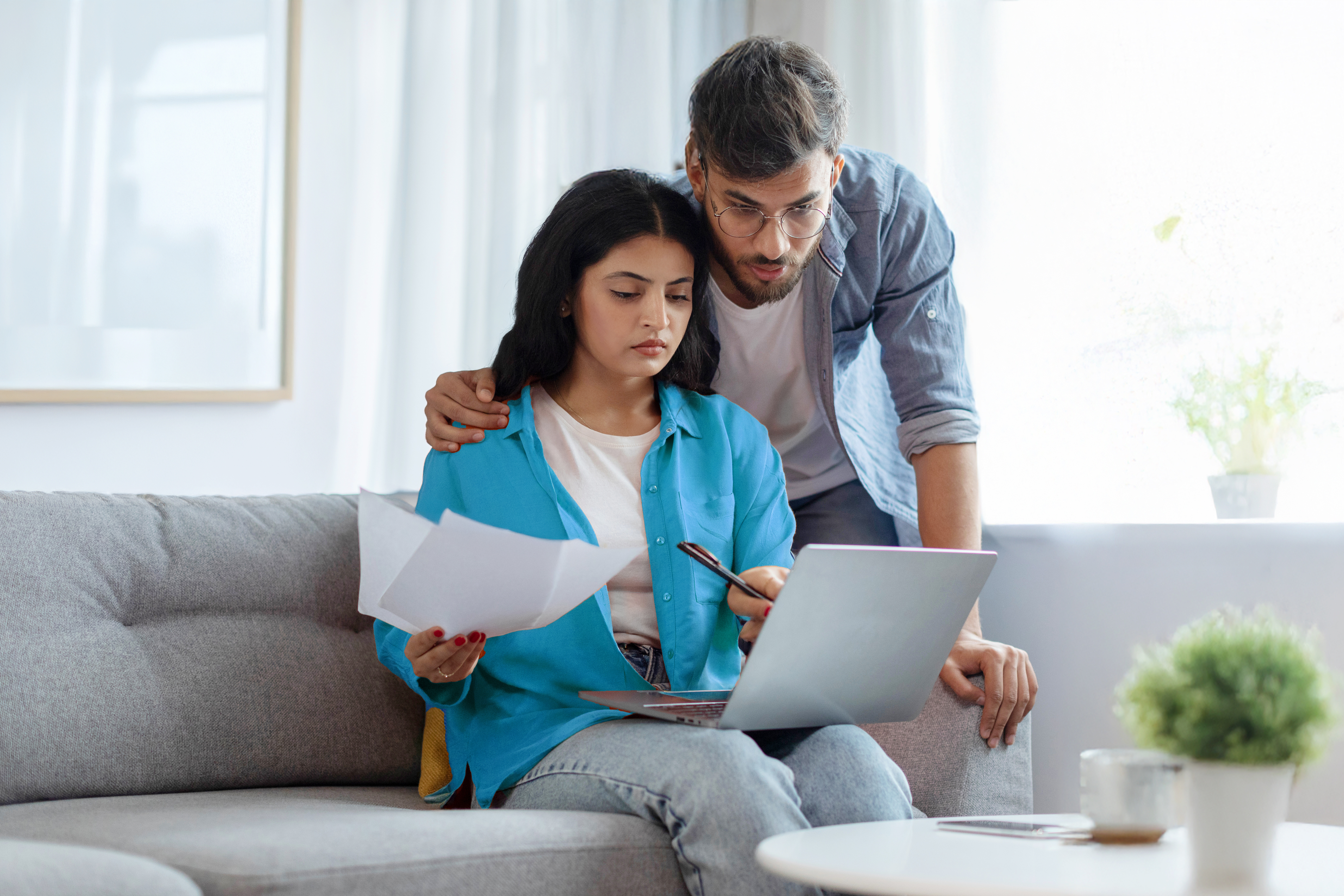 Household papers problem. Indian couple looking at laptop and holding documents, reading bills and correspondence, home interior